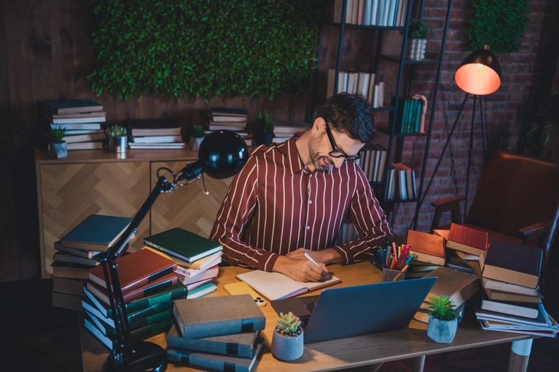 A young man works and studies surrounded by books in a warm, stylish home office. He actively writes on paper while referencing materials under soft lighting, showcasing focus and comfort.