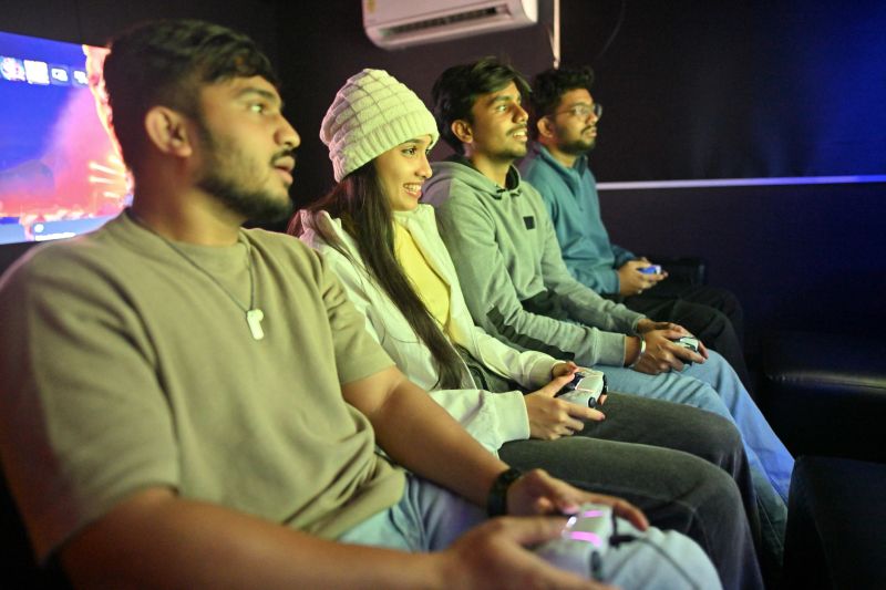 A fun and energetic scene inside a gaming parlor, featuring a group of young men and a young woman, each holding a controller as they play video games together. The group is focused on their screens, with expressions of concentration and excitement. This moment captures the joy of social gaming and friendly competition in a vibrant setting.