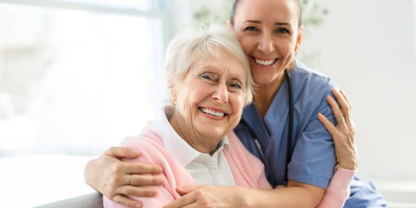 A nurse warmly embraces a smiling elderly woman in a bright room.