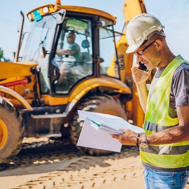 Construction worker in safety gear checks documents and talks on phone at site with heavy machinery.