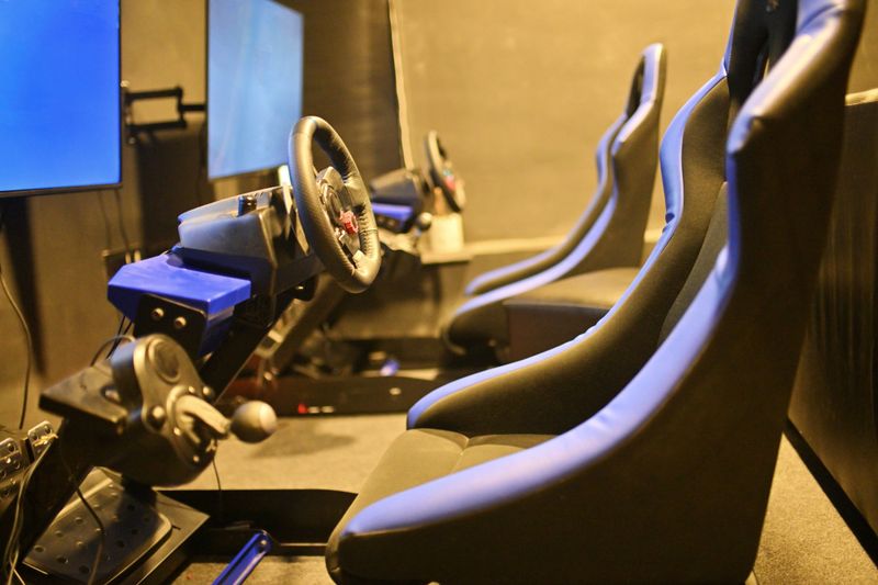 A fully equipped car racing gaming station, unoccupied and ready for the next player. The setup features a large screen displaying a racing game,  a steering wheel, and responsive pedals for an immersive driving experience.