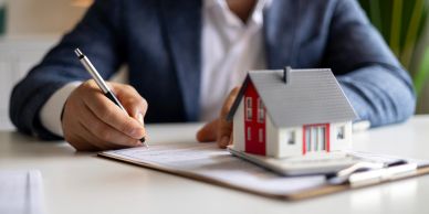 Person signing real estate documents beside a house model.