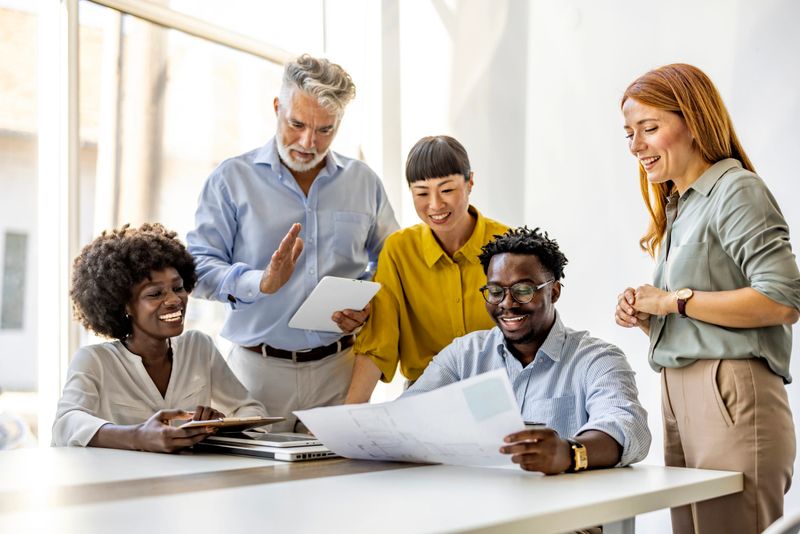 A diverse group of professionals collaborating around a table in a modern office. They are discussing documents with positivity and teamwork. The atmosphere is productive and inclusive, emphasizing collaboration.