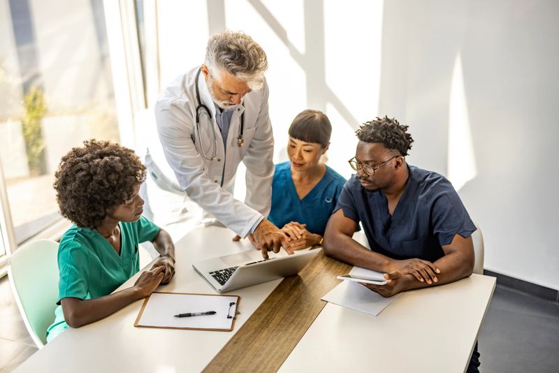 A diverse group of healthcare professionals, including a senior doctor and nurses, discussing patient diagnosis together in a bright meeting room. The team is focused on a laptop screen.