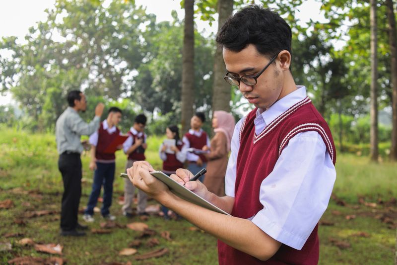 Portrait Of Student With Deaf Holding Clipboard And Writing Notes Against Group Of Students Exploring The Environment Background