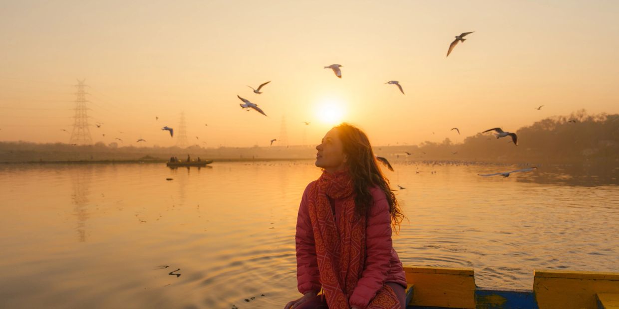 Woman enjoying sunrise by the river with birds flying.