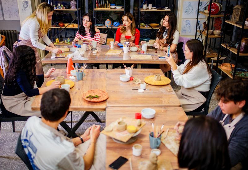 A pottery instructor demonstrates a clay modeling technique to a group of engaged students in a creative workshop setting. The participants, mostly women, sit around a large wooden table filled with sculpting tools, clay pieces, and ceramic items. The studio’s artistic ambiance, complete with shelves of pottery and design sketches, highlights the collaborative and educational atmosphere of the class.