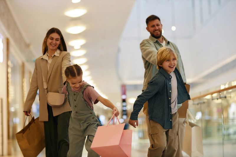Wide shot of happy children with gift bags holding parents hands while running in shopping mall