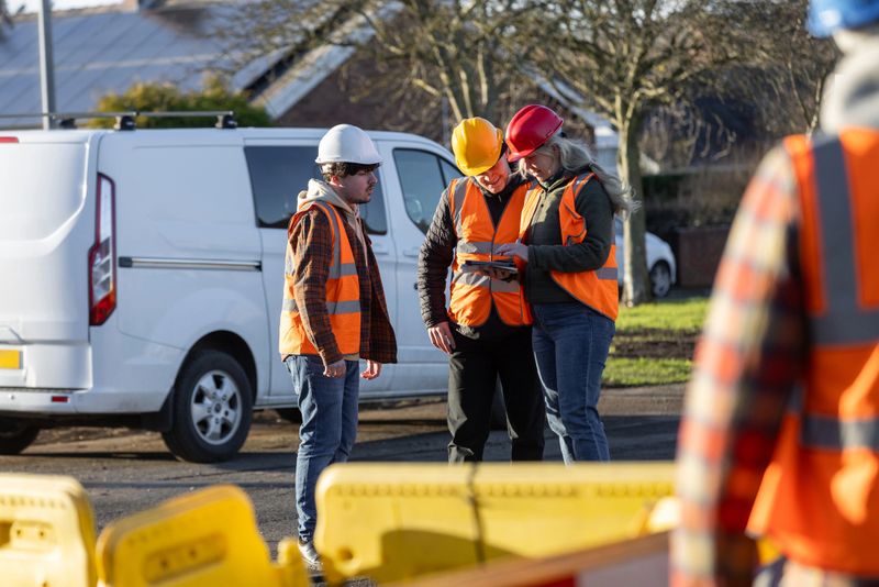 Wide-view of three construction workers engaged in a discussion at an outdoor worksite in Seghill, North East England. They're wearing safety gear, including hard hats and high-visibility vests, while standing together on site near construction barriers and their work van. One construction worker is using a digital tablet.

Videos similar to this scenario are available.