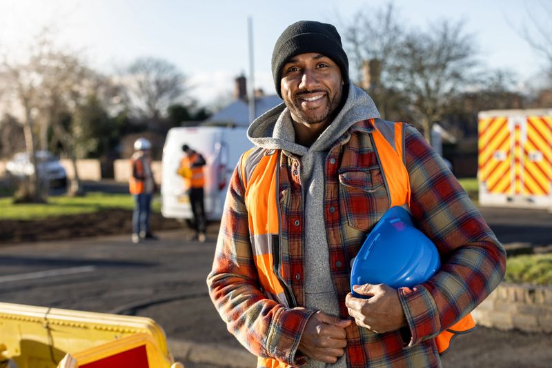 Front-view portrait of a mid adult male construction worker at an outdoor worksite in Seghill, North East England. He is wearing safety gear, including a high-visibility vest and holding a hard hat, while standing on site near construction barriers and his work van. He is deciding his next plan of action while looking at the camera.