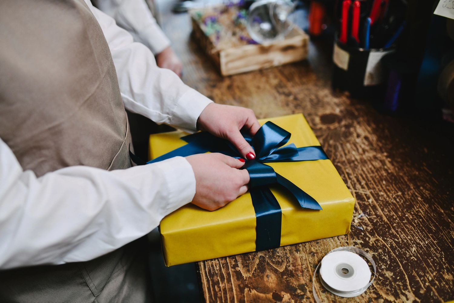 Hands tying a blue ribbon on a yellow gift box on a wooden table.