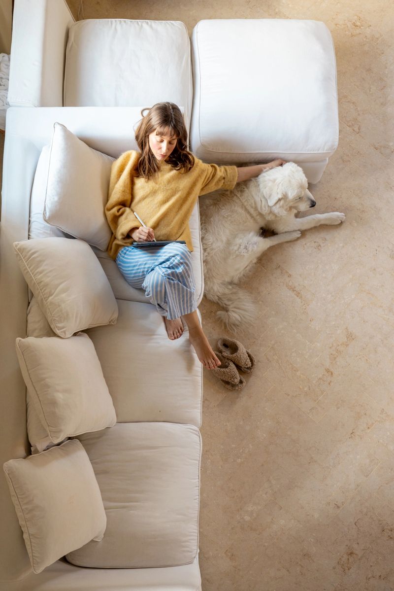 A young woman in a warm yellow sweater and striped pajama pants relaxes on a white sofa, gently petting her fluffy large dog. The scene exudes warmth, comfort, and tranquility in a modern home setting