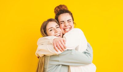 Two young women happily hugging each other against a bright yellow background.