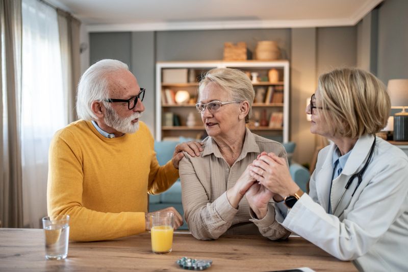 Doctor is holding the hands of a senior woman, comforting her while her husband is holding her shoulder, providing support during a difficult time at home