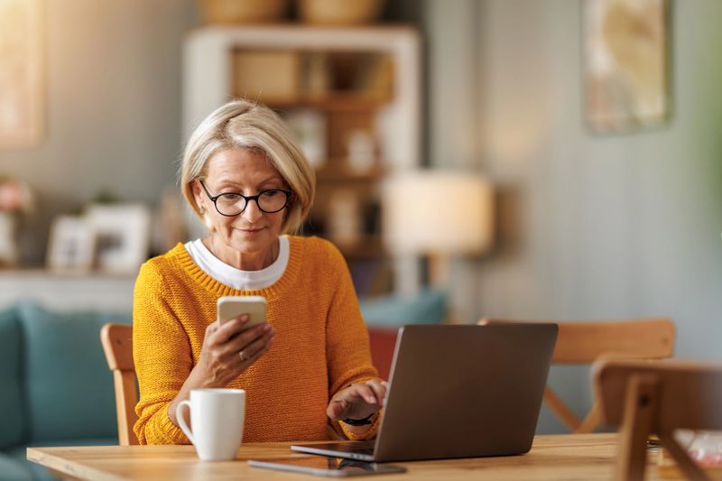 A woman in a cozy orange sweater sits at a wooden table, using a smartphone in one hand and typing on a laptop with the other. A cup of coffee is nearby, creating a warm atmosphere.