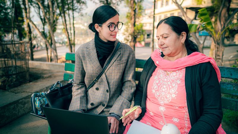 A young woman in professional attire and glasses sits on a park bench with a senior woman wearing a traditional outfit. They engage in a learning or work session sitting on the park bench. The young woman uses a laptop computer, and the senior woman holds a notebook, pen, and coffee cup. The scene shows a knowledge exchange between generations in a peaceful outdoor setting with trees and natural light.