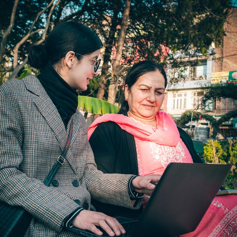 A young woman in professional attire and glasses sits on a park bench with a senior woman wearing a traditional outfit. They engage in a learning or work session sitting on the park bench. The young woman uses a laptop computer, and the senior woman holds a notebook. The scene shows a knowledge exchange between generations in a peaceful outdoor setting with trees and natural light.