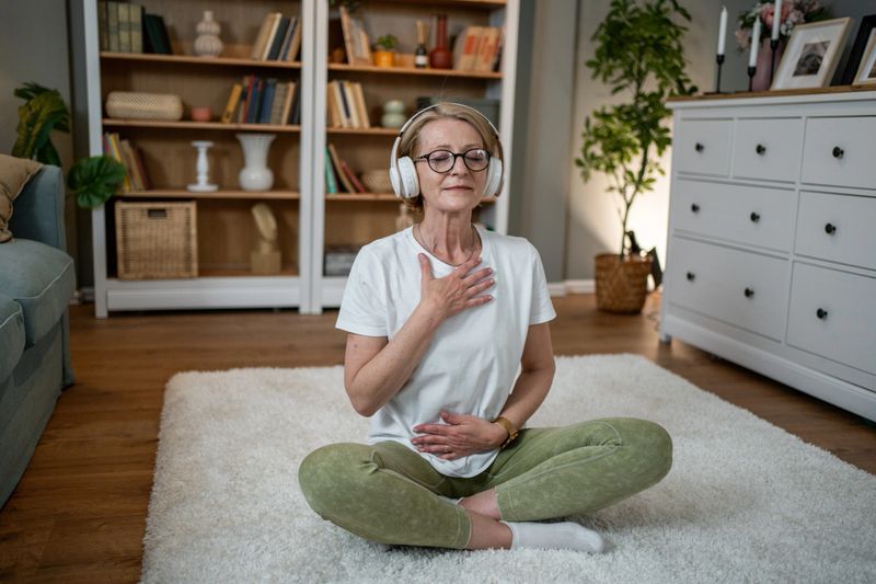 Mature woman sitting in lotus position on a white rug, wearing headphones and practicing meditation at home, with her eyes closed and hands on her chest and belly.