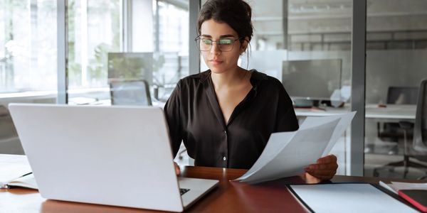 Woman in glasses working on laptop while holding papers in a modern office.