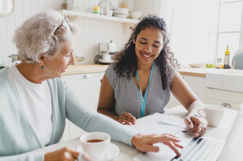 Side view of black female volunteer helping senior caucasian lady to improve her computer skills, teaching her to use wireless internet, laptop, browsing social media, web pages, checking e-mail