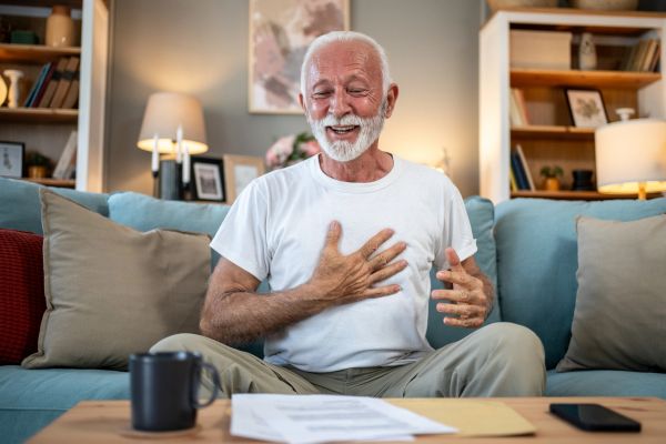 Elderly man smiling on couch with papers and mug on table.