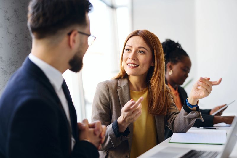 Businesswoman explaining new project to businessman during a meeting in a modern office, with another colleague using a tablet in the background