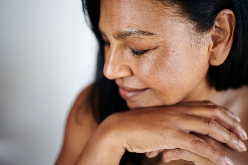 A serene mature woman of South Asian ethnicity sits with her eyes closed, hands gently resting on her chin, radiating calm and contentment in soft natural light.
