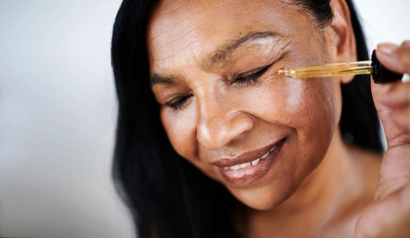 Mature woman with radiant skin smiling while applying facial oil using a dropper.