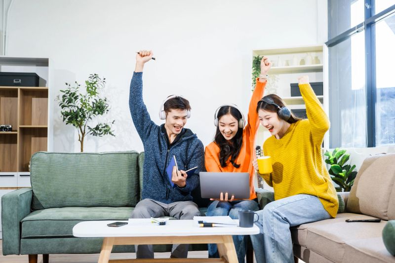 Three Asian young adult students sit in university living room, discussing their study abroad experiences, academic research, preparing thesis report, planning future global education opportunities.
