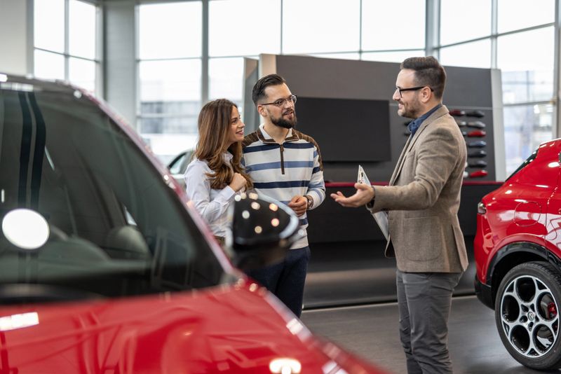 Professional car salesman is holding a tablet and showing a new red car to a young couple in a modern car dealership, discussing the features and benefits of the vehicle