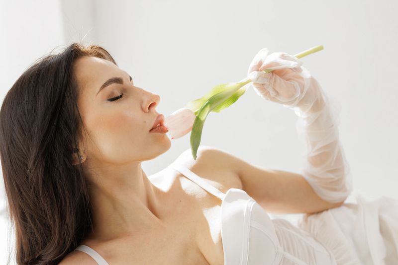 Elegant woman in white lingerie with delicate gloves holding a pink tulip near lips in soft natural light