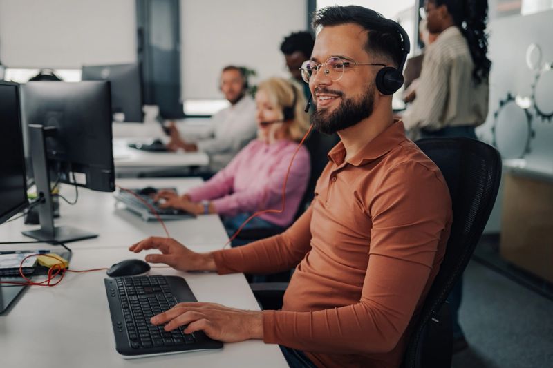 Smiling male call center agent wearing a headset types on a keyboard while working at a computer in a busy office with diverse colleagues assisting customers