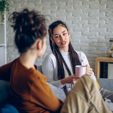 Two women engaged in a cozy conversation indoors, one holding a pink mug.