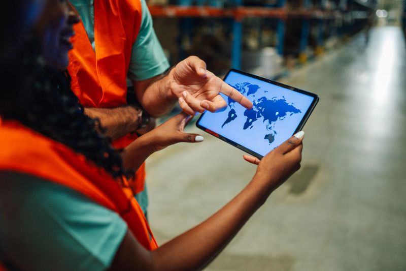 Two warehouse workers are using a digital tablet showing a world map, discussing logistics, delivery, and global shipping strategies in a modern storage facility