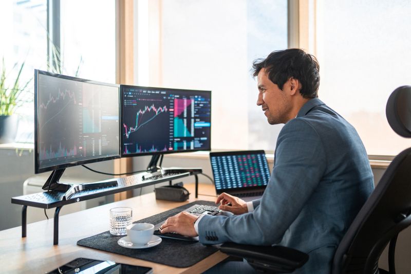 Profile view of mid-adult Asian financial trader working at desk with multiple monitors displaying market data. Natural light illuminates modern workspace with professional setup.