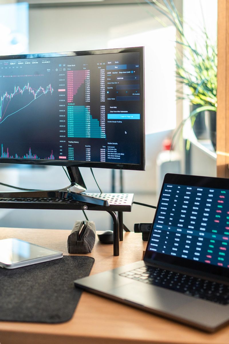 Professional trading setup with desktop monitors and laptops displaying stock market data in a bright office environment with natural lighting.