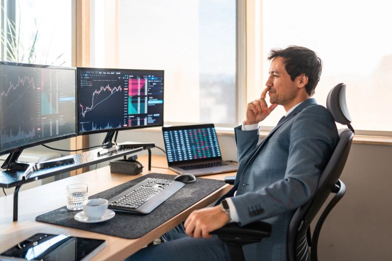 Profile view of mid-adult Asian financial trader working at desk with multiple monitors displaying market data. Natural light illuminates modern workspace with professional setup.