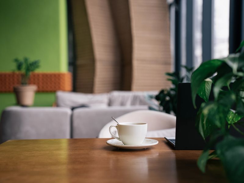 Front view of warm cup of coffee rests on a wooden table, accompanied by a spoon. Interior of modern cafe with comfortable seating and full of bright greenery. White cup of americano near laptop.