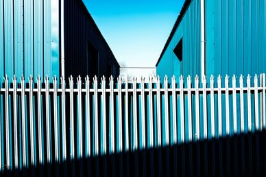 Metal fence in front of two blue industrial buildings under clear sky.