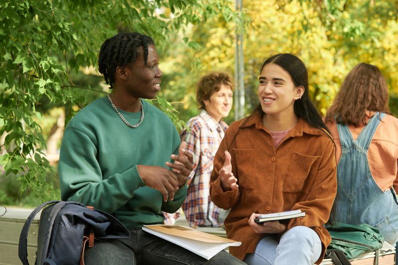 Group of students enjoying outdoor study session under trees while conversing and smiling. Focusing on blended learning environment with lush greenery in background