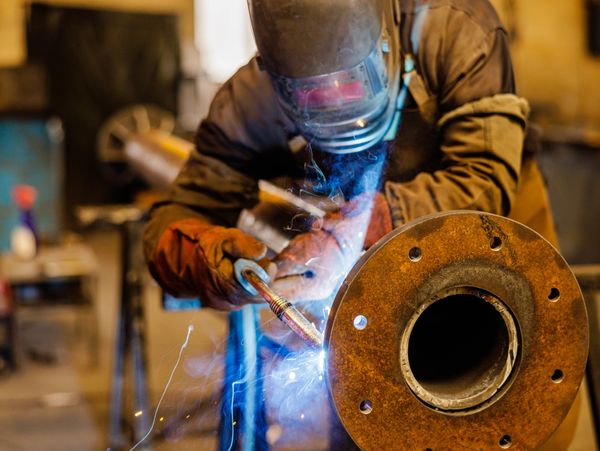 Welder in protective gear welding a large rusty metal pipe indoors.