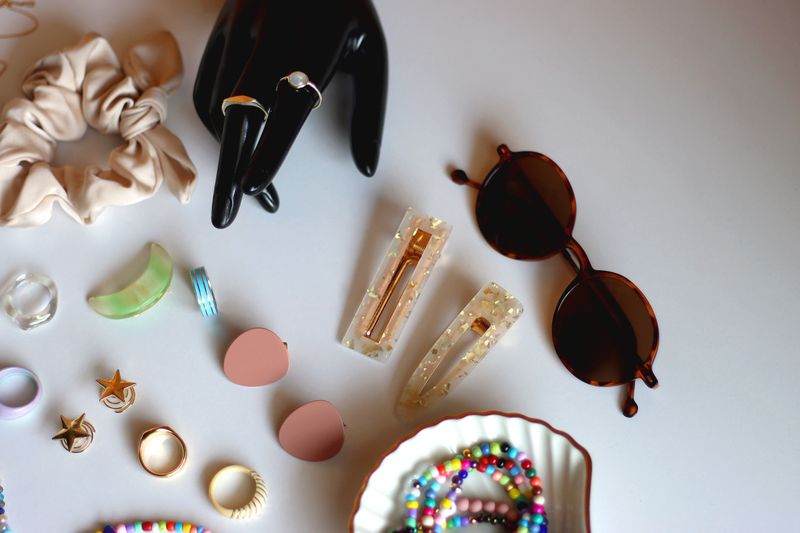 Hand figurine, sunglasses, gold jewelry, colorful bead jewelry, statement rings and various hair accessories on the white table. Selective focus.