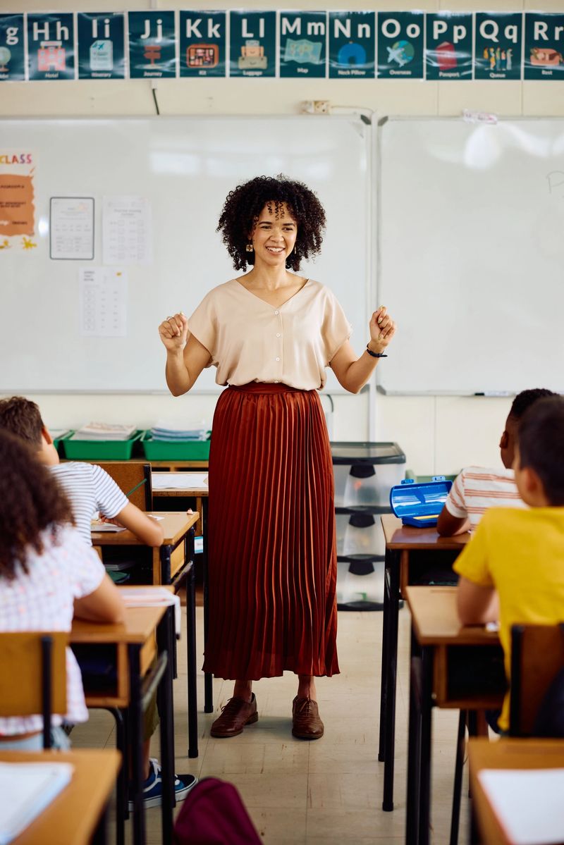 School children sitting at desks, listening attentively to young cheerful female teacher talking and gesturing in class