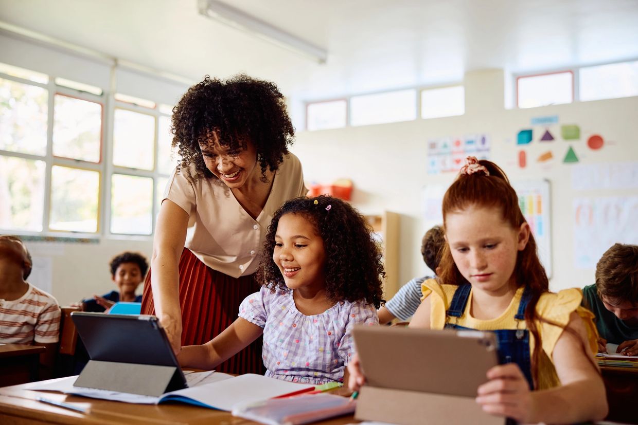 Teacher smiling while helping a student with a tablet in class.