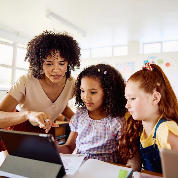 Teacher guiding two young girls using a tablet in a classroom.