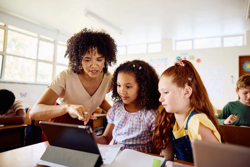 School teacher helping and guiding schoolgirls sitting at desk using digital tablet in classroom