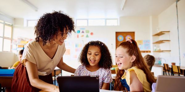 Teacher and two young girls happily looking at a laptop in a classroom.