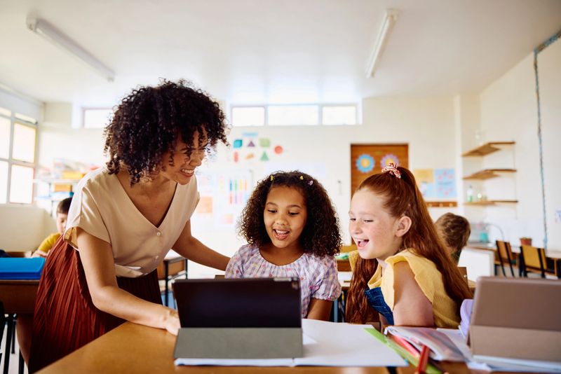 School teacher smiling to schoolgirls sitting at desk using digital tablet, explaining and laughing