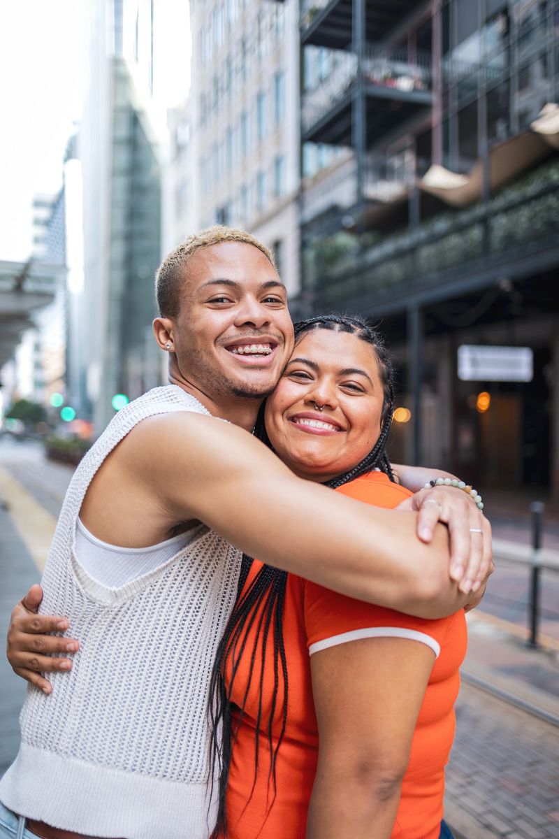 Two friends are embracing and smiling warmly, showcasing genuine happiness outdoors in an urban setting. The image captures a candid and heartfelt connection in a lively city environment.