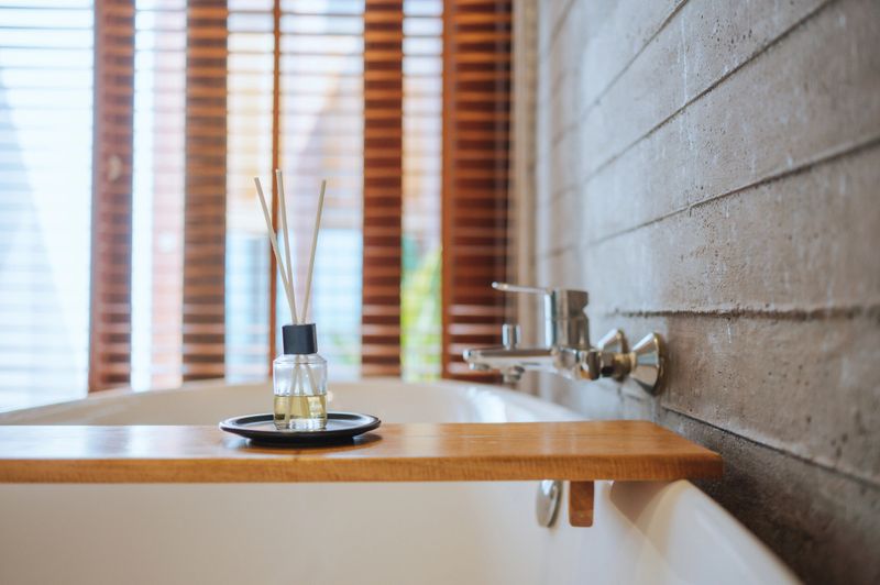 A glass reed diffuser with black sticks placed on a wooden counter near a modern bathroom sink, creating a relaxing and elegant home spa ambiance.